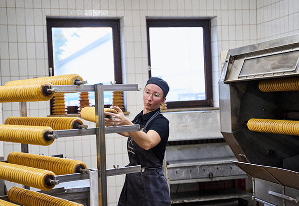 Eine Baeckerin backt Baumkuchen auf rotierenden Rollen in einer Backstube.
