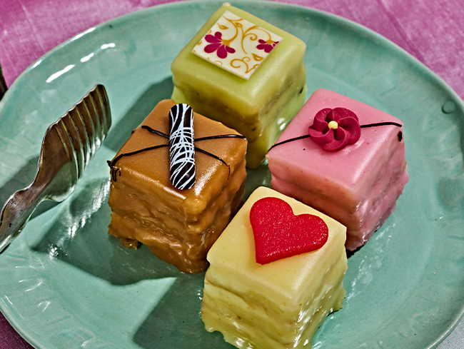 Vier farbige Petit Fours mit Deko auf gruenem Teller, Gabel links daneben, rosa Hintergrund.