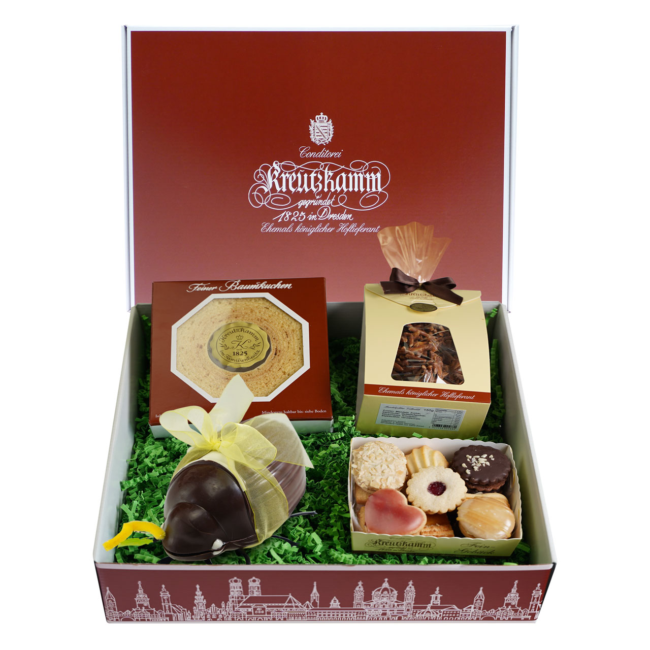 Maigeschenkpaket-01 Geschenkbox mit Logo der Konditorei Kreutzkamm, gefuellt mit Gebaeck und Pralinen, umgeben von gruenem Verpackungsmaterial.