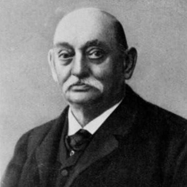 Ein aelterer Mann mit Schnurrbart traegt Anzug, sitzt und schaut in die Kamera, neutraler Hintergrund.
