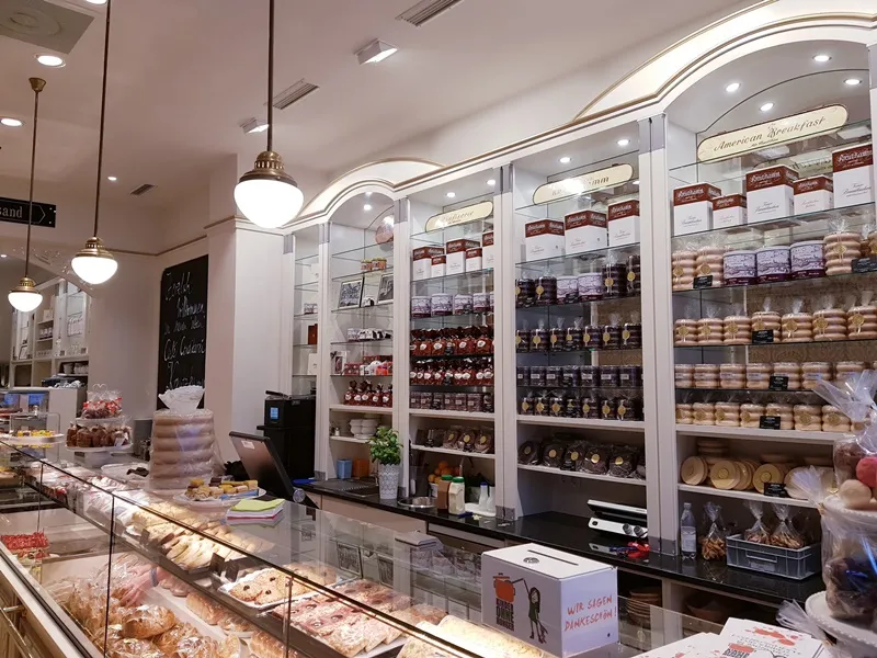 Konditorei mit Gebaeckvitrine. Verkaufsregale gefuellt mit Marmeladen- und Keksdosen. Im Hinter- grund steht "American Breakfast".
Text:
Wir sagen Dankeschoen