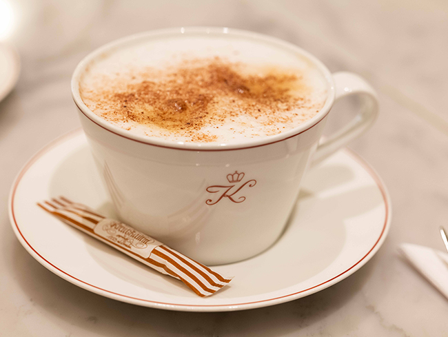 Eine Tasse Milchschaumkaffee steht auf einer Untertasse, neben einer verpackten Zuckerruetung. Auf der Tasse befindet sich der Buchstabe "K" mit einer Krone.