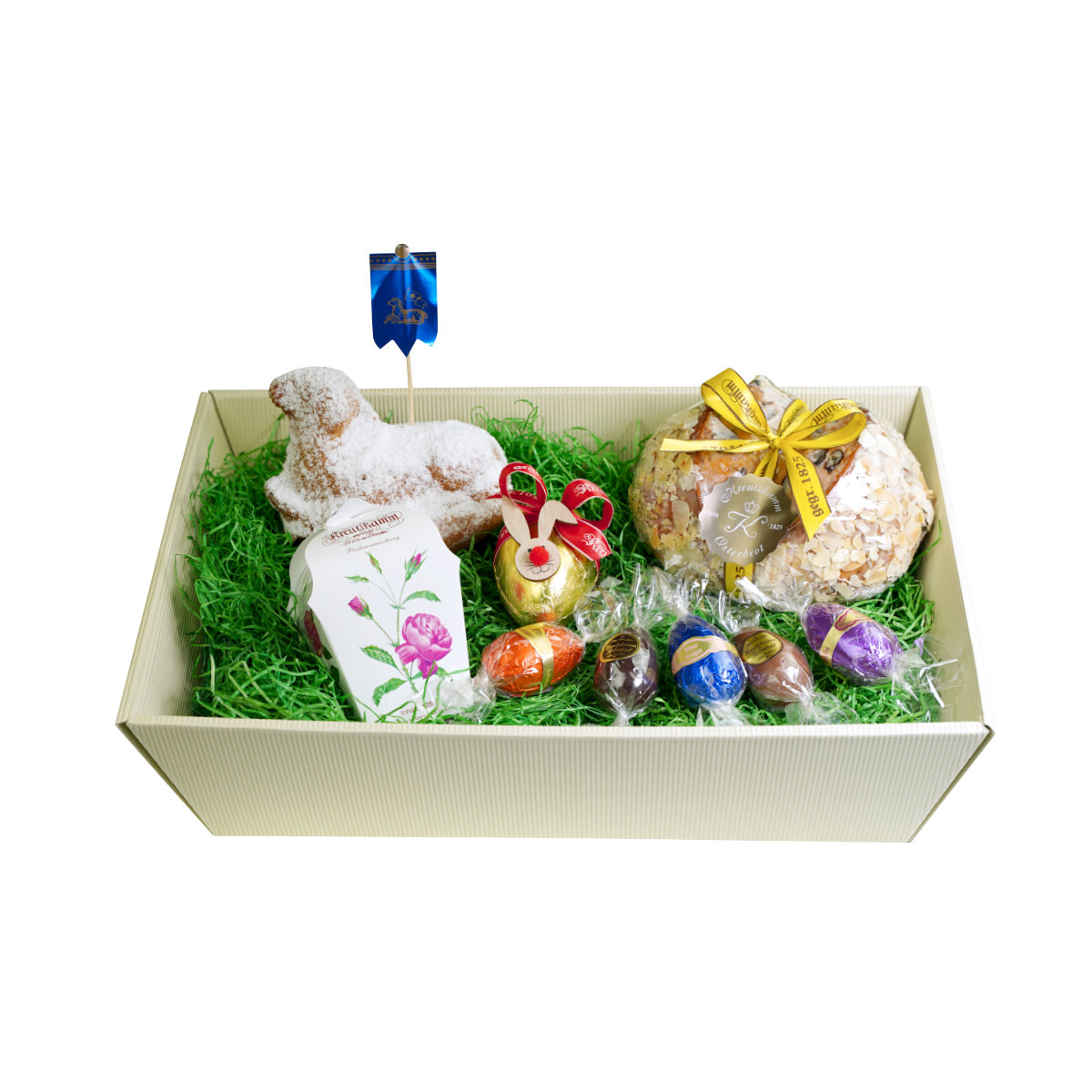 Ein Osterkorb mit einem Gebaecklamm, Schokoladeneiern, Gebaeck und einer Goldverpackung, umgeben von gruenem Kunststoffgras. Text: "Backhaus Kreutzkamm".