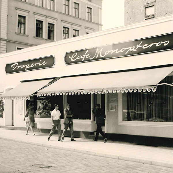 Mehrere Personen spazieren vor Drogerie und Cafe Monopteros auf einer staedtischen Strasse.