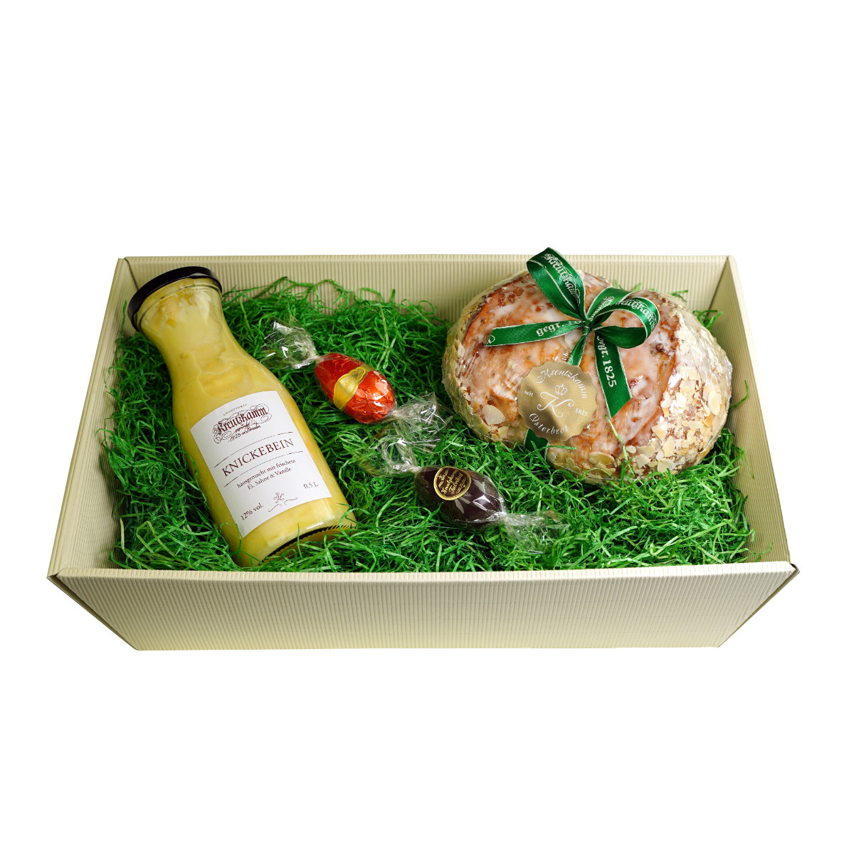 Geschenkbox mit einer Flasche Knickebein-Likoer, einer glasierten Brotkugel und kleinen Schokoladen in gruenem Gras. Text auf der Flasche: "Kreutzkamm" und "Knickebein".