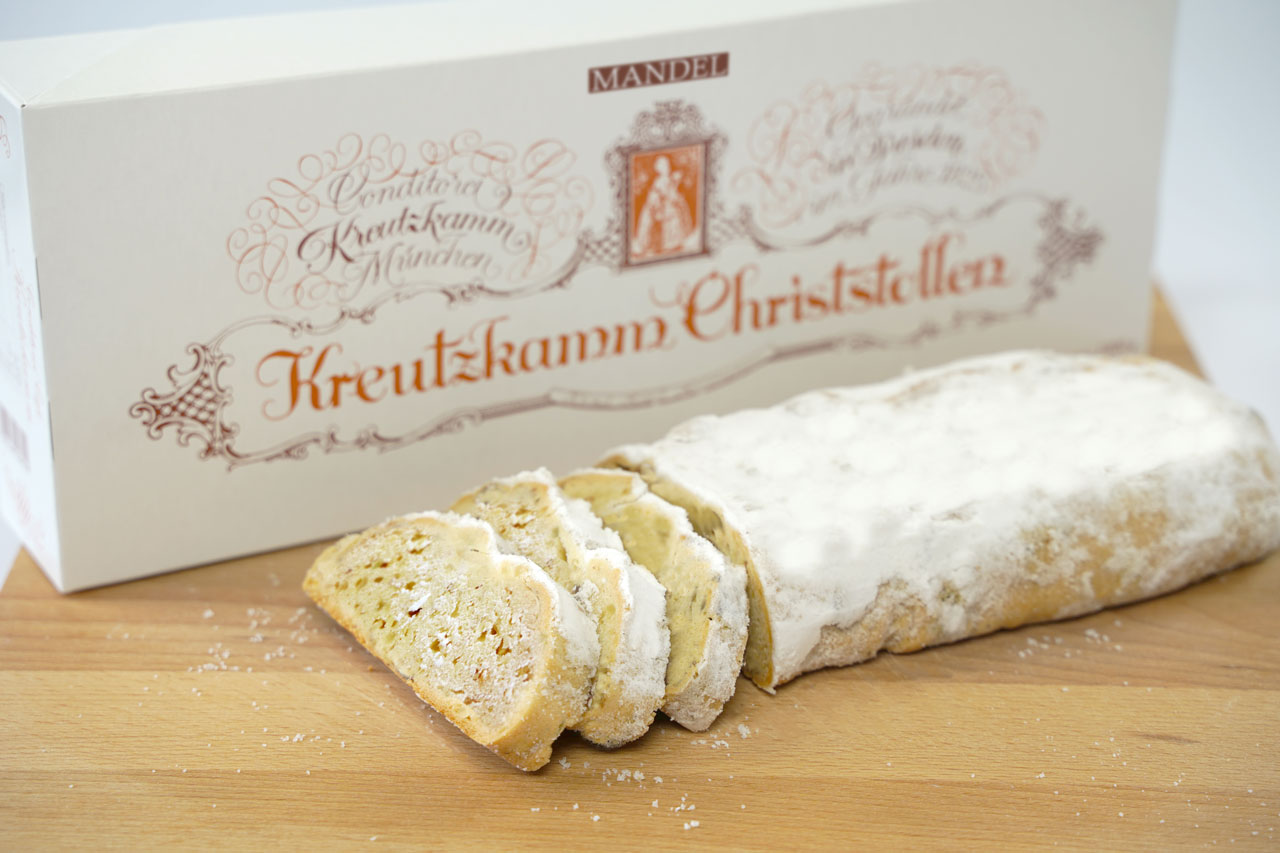 Ein angeschnittener Christstollen mit Puderzucker steht auf einem Holzbrett. Dahinter eine Verpackung mit der Aufschrift "Konditorei Kreutzkamm Muenchen, Kreutzkamm Christstollen".