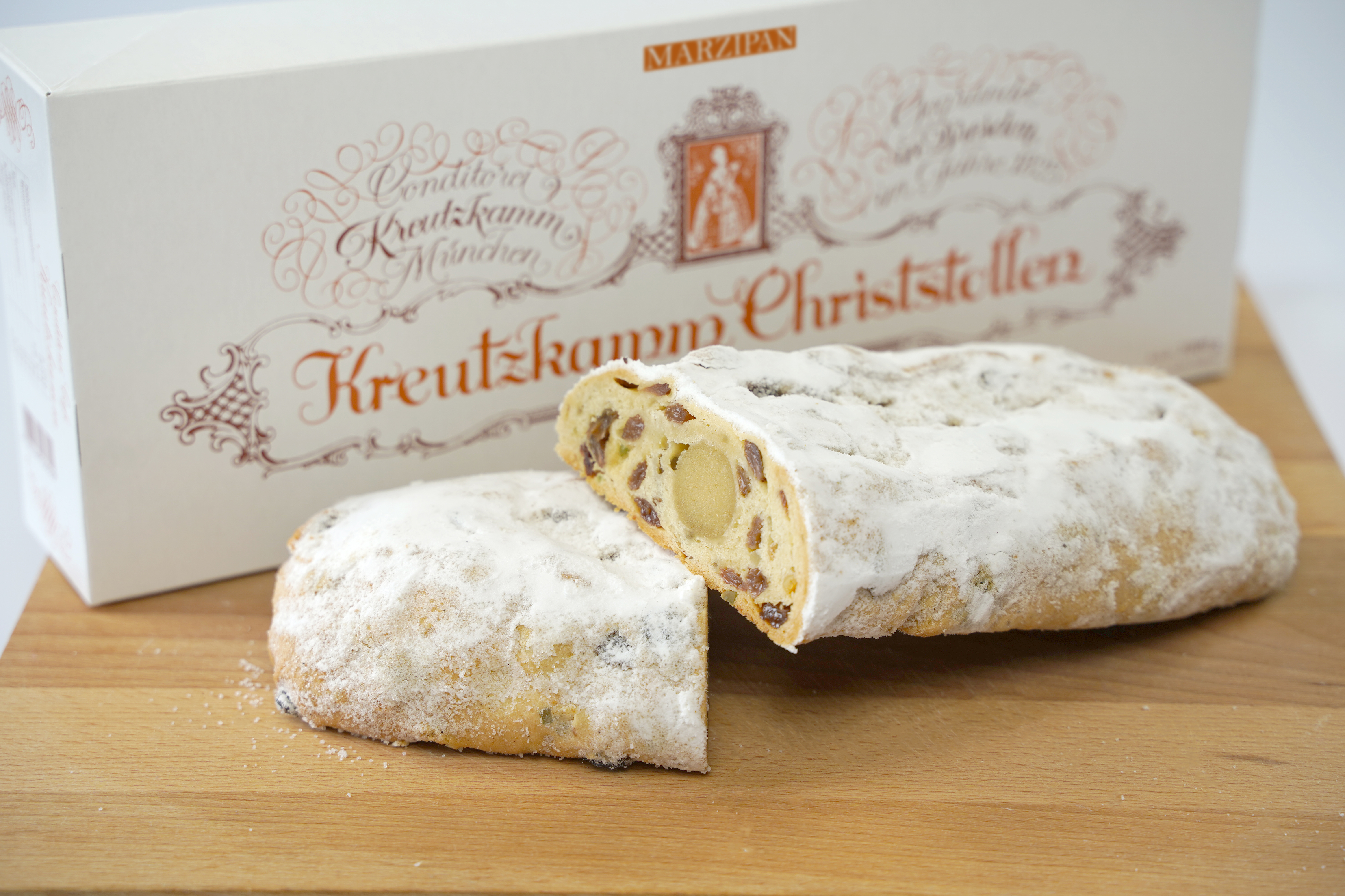 04-Marzipantstollen Stollen, angeschnitten, auf Holzbrett. Hintergrund: Verpackung mit Text: Konditorei Kreutzkamm Muenchen, Kreutzkamm Christstollen, MARZIPAN.