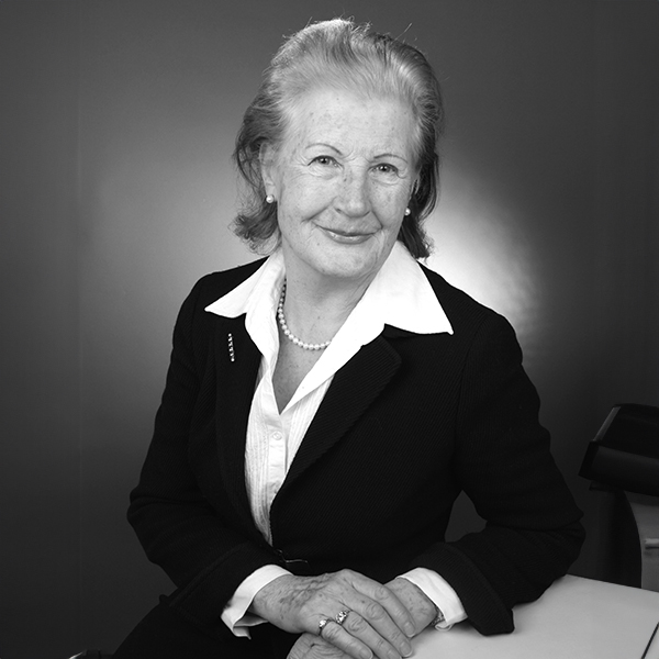 Eine Frau sitzt laechelnd an einem Tisch, traegt einen Blazer und eine Perlenkette, grauer Hintergrund.