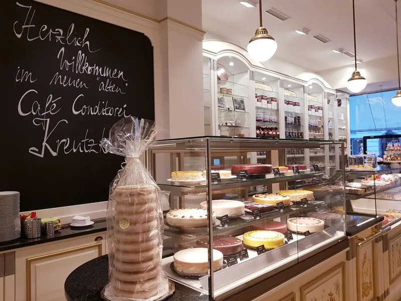 Schwarzes Brett mit der Aufschrift: "Herzlich willkommen im neuen alten Cafe Conditorei Kreutzkamm" links. Kuchenvitrine rechts.