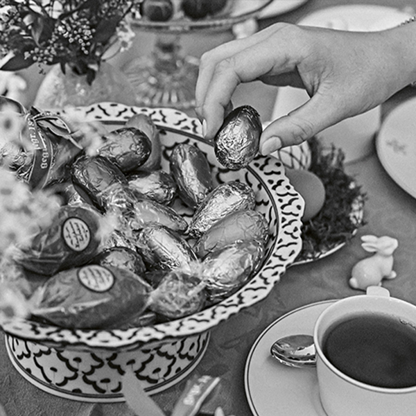 Hand greift nach pralinenverpackten Eiern aus dekorativer Schuessel auf einem Tisch mit Tee.