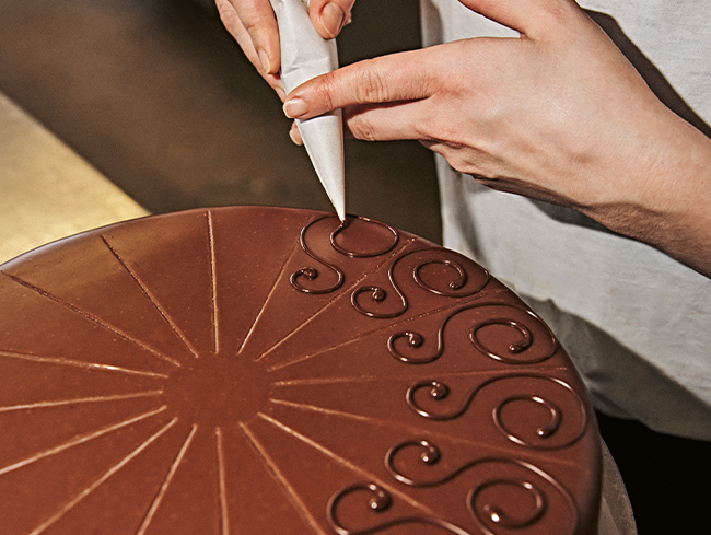 Person verziert Schokoladenkuchen mit Zuckerguss, helle Umgebung.