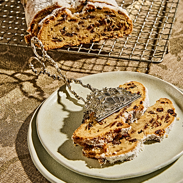 Zwei Scheiben Stollen auf einem Teller, dekorative Zange, Gitter mit restlichem Stollen im Hintergrund